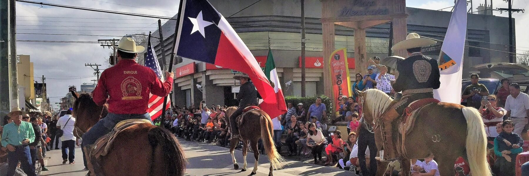 A parade along the border (frontera) with participants in cowboy hats on horseback. The main rider in focus is holding a Texas flag. Viewers cheer from along the edge of the street in a dense crowd.