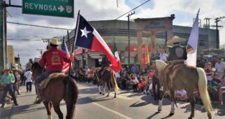 A parade along the border (frontera) with participants in cowbow hats on horseback. The main rider in focus is holding a Texas flag. Viewers cheer from along the edge of the street in a dense crowd.