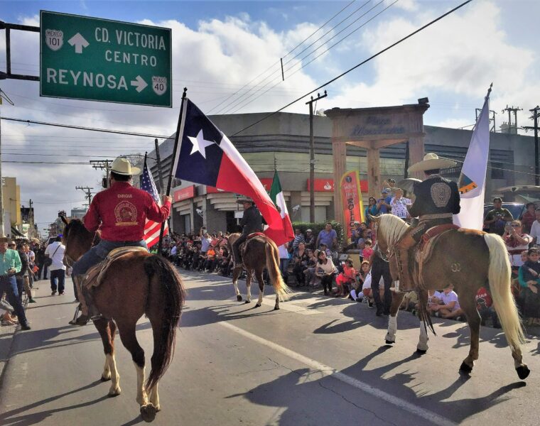 A parade along the border (frontera) with participants in cowboy hats on horseback. The main rider in focus is holding a Texas flag. Viewers cheer from along the edge of the street in a dense crowd.